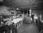 Men sit on stools at a counter and workers stand nearby in an unidentified cafe by Basil Clemons 1887-1964