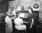 A group of adult and young women gathered around a piano by Basil Clemons 1887-1964