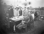 A group of men and women sitting at a table outdoors, eating by Basil Clemons 1887-1964