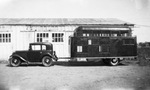 A two-door car with a large covered trailer attached at the rear parked outside a garage by Basil Clemons 1887-1964