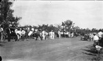 People stand on both sides of a road as a race car marked #1 driven by a man wearing goggles comes into view by Basil Clemons 1887-1964