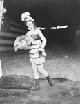 Female circus performer wearing a ruffled and feathered costume stands under a large circus tent near a large American flag, Breckenridge, Texas by Basil Clemons 1887-1964