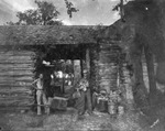 Two men stand beside a log cabin; one man holds deer antlers; location not identified by Basil Clemons 1887-1964