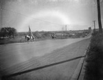 Riders in Native American dress riding bareback on horses and carrying the U.S. flag as they come out of downtown Breckenridge, Texas by Basil Clemons 1887-1964