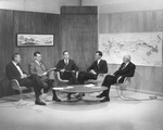 Five men sitting around small round table with microphones in hand