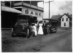 Three people standing on unpaved street beside cars in front of a bank