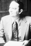 Photograph of a man seated at a desk. The man is wearing a sport jacket and a wide paisley-patterned tie by Squire Haskins Photography Inc.