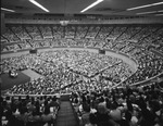 Large auditoriam filled to capacity with men and women. A man is speaking on a raised platform. Many of the attendees are holding identical but unidentifiable books by Squire Haskins Photography Inc.